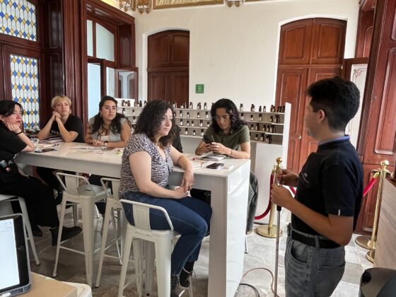 Foto de un hombre con discapacidad visual impartiendo una clase a un Equipo de guías del museo del perfume. Están en un salón de un edificio antiguo, con muchos detalles esculpidos en el techo.